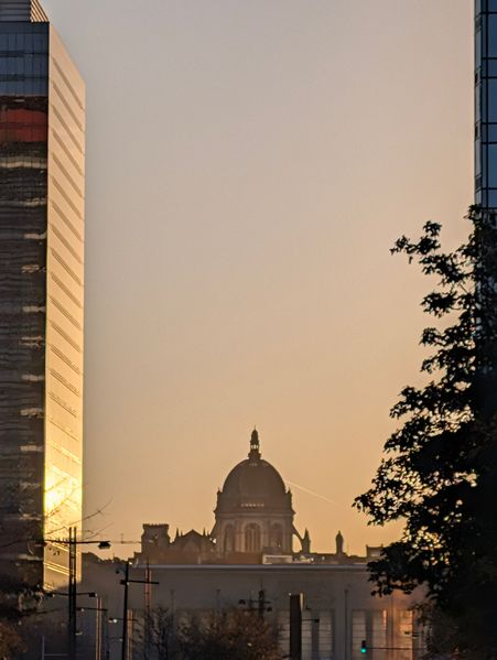 A domed building is framed by two tall structures and silhouetted trees under a warm, glowing sky.